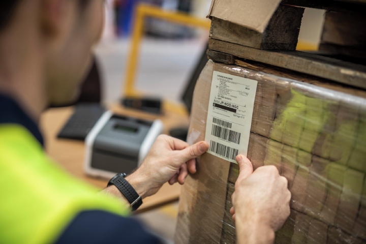  Ein Arbeiter beklebt ein Paket mit einem Barcode. Im Hintergrund ist verschwommen der Etikettendrucker zu sehen. 