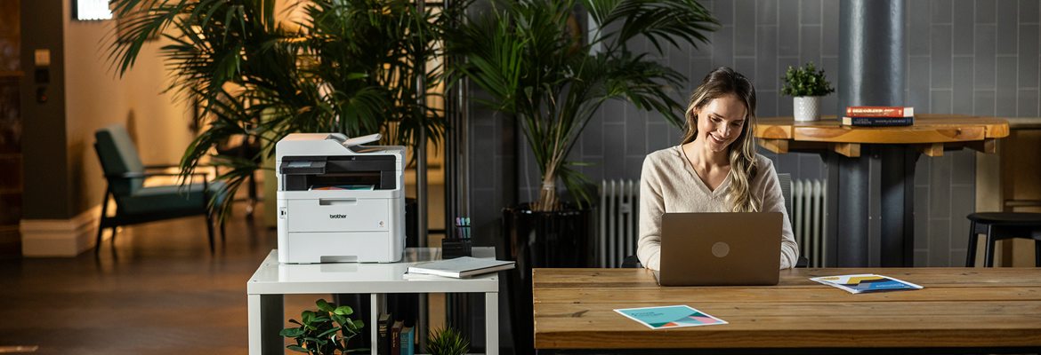 Eine Frau sitzt im Büro am Schreibtisch. Brother Drucker im Hintergrund.