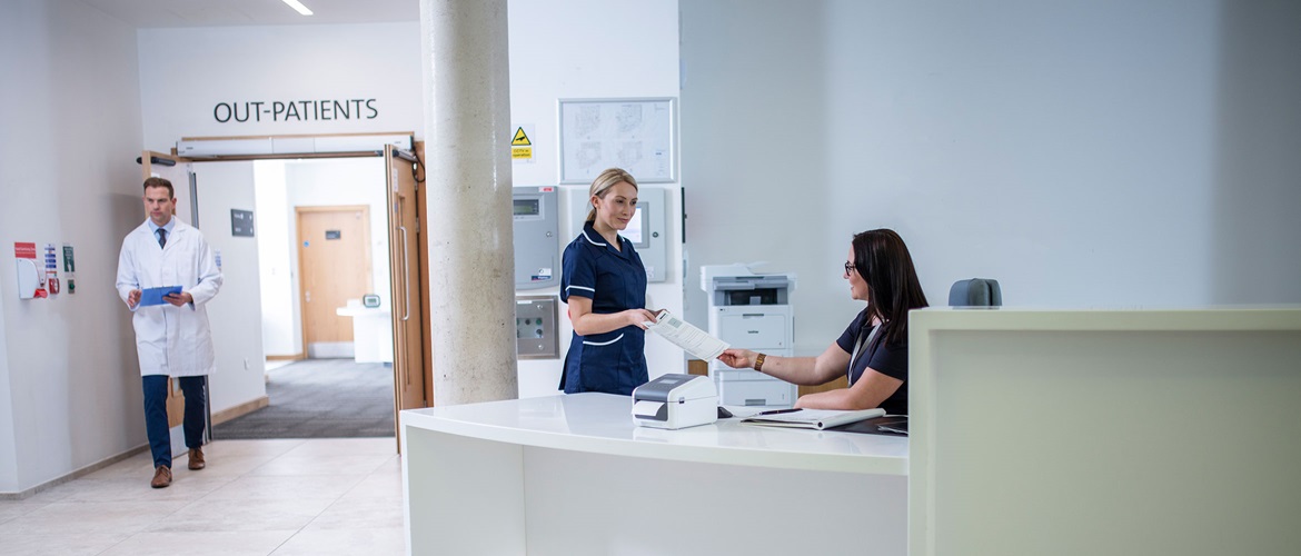 Artzhelferin am Schreibtisch, Während Einer Krankensschwwester und der Arzt im Hintergrunder Arbeiten.