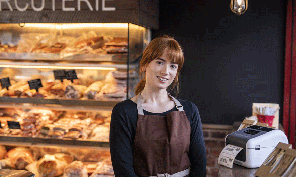 Eine Mitarbeiterin in einer Bäckerei steht neben einem Etikettendrucker von Brother, der gerade ein Preisetikett ausdruckt