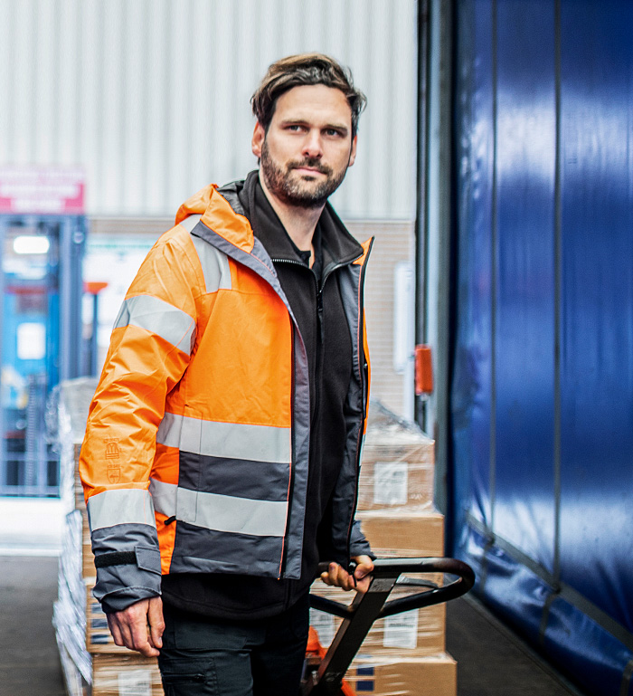 MANN MIT ORANGEEFARBENER WISSCHUTZJAKE ZIEHT HUBWAGEN MIT PALETTE UND KISTEN IM LKW MIT BLAUEN SEINEN
