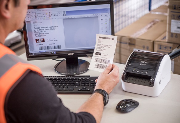 Arbeiter sitzt vor dem Laptop und vor dem QL-1100 im Logistikzentrum und druckt Versandetiketten aus 