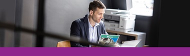 Ein Mann sitzt im Büro. Brother Drucker, der über Managed Print Service verfügt, im Hintergrund.