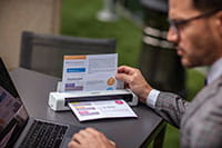 Man wearing grey suite with glasses, sat outdoors using laptop and Brother DSmobile DS740D portable document scanner with colour A4 document