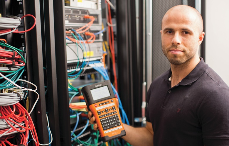 Der männliche Netzwerktechniker steht vor einem ordentlich markierten Netzkabinett, während er ein Beschriftungsgerät von Brother hält