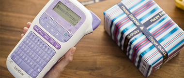 	 Eine Hand hält ein lilafarbenes P-touch-Etikettiergerät, im Hintergrund ein Tisch mit einer als Geschenk verpackten Schachtel, die mit einem bedruckten Band verschnürt ist