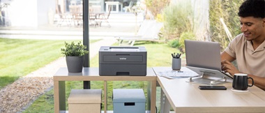Mann sitzt an einem organisierten Schreibtisch und schaut auf einen Laptop neben einem Drucker und Kisten auf einem Beistelltisch mit einem Garten im Hintergrund