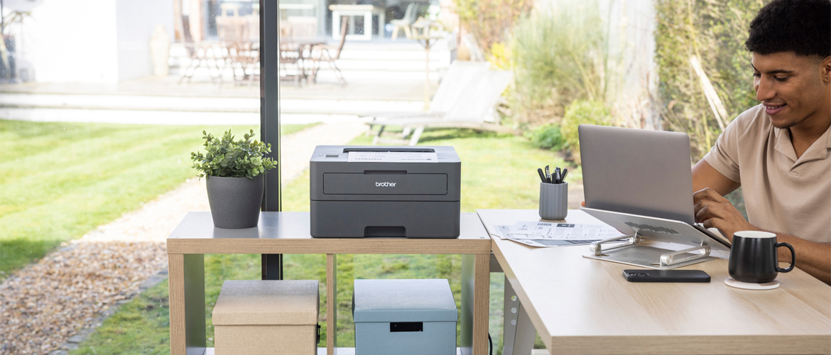 Mann sitzt an einem organisierten Schreibtisch und schaut auf einen Laptop neben einem Drucker und Kisten auf einem Beistelltisch mit einem Garten im Hintergrund