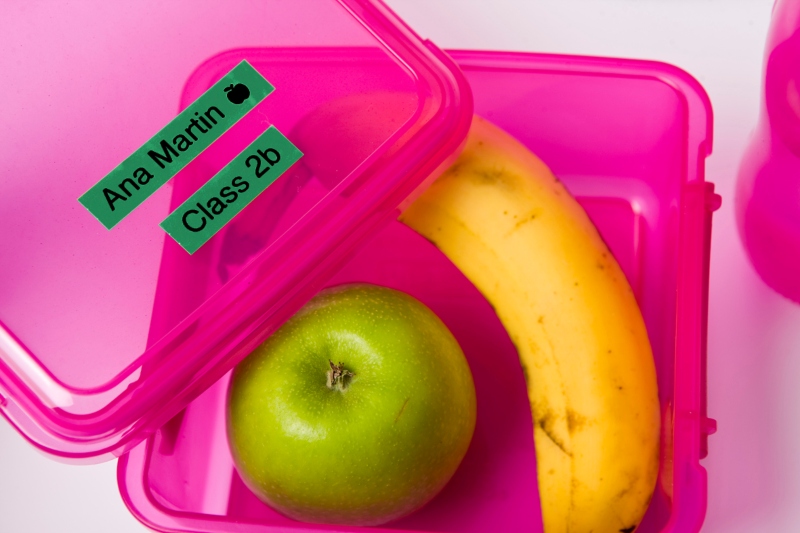 Geöffnete rosa Lunchbox mit einem Apfel und einer Banane darin und einem grünen Etikett oben drauf