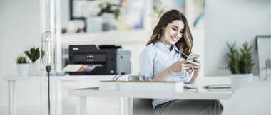 Eine Frau sitzt an ihrem Schreibtisch und schaut auf ihr Smartphone. Im Hintergrund ist ein Drucker auf einer Kommode zu sehen.