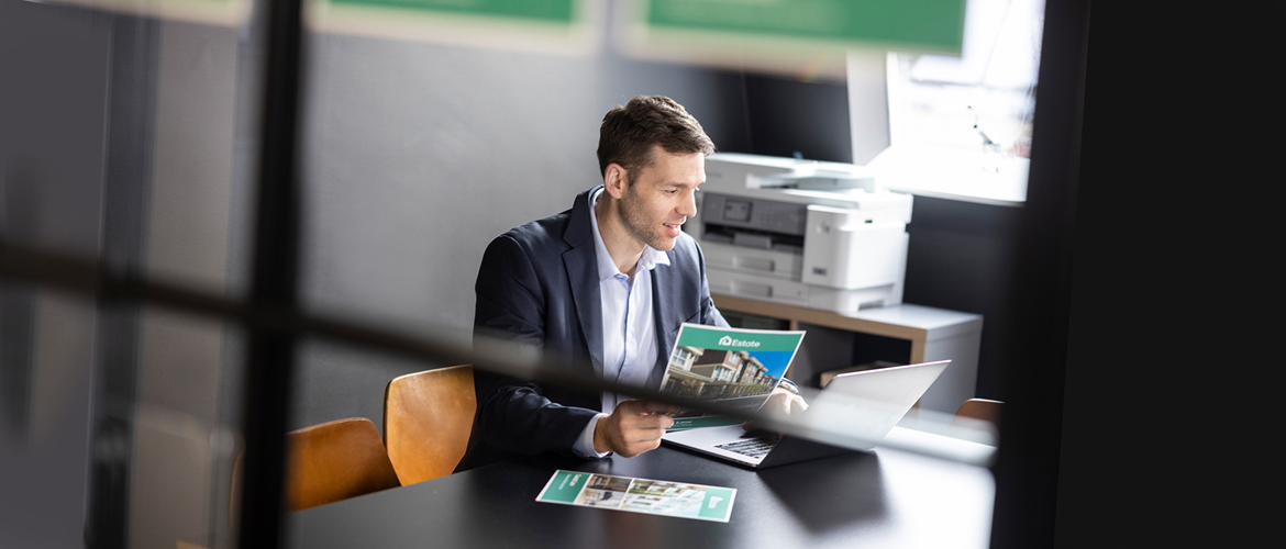 Ein Mann sitzt an einem Schreibtisch und hält ein Blatt Papier in der Hand. Brother Drucker im Hintergrund.