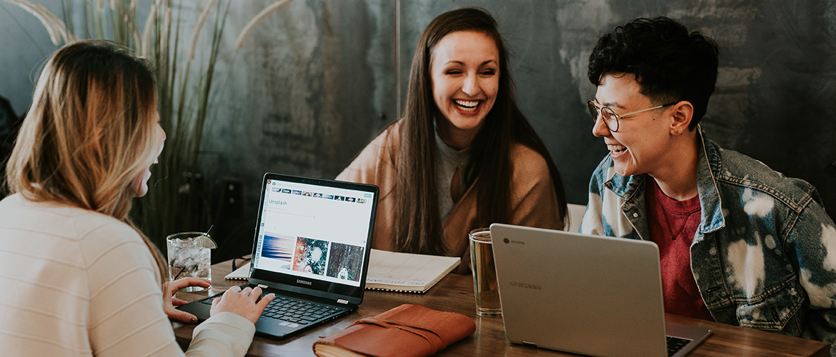Drei Frauen sitzen am Tisch mit Laptops und lachen.
