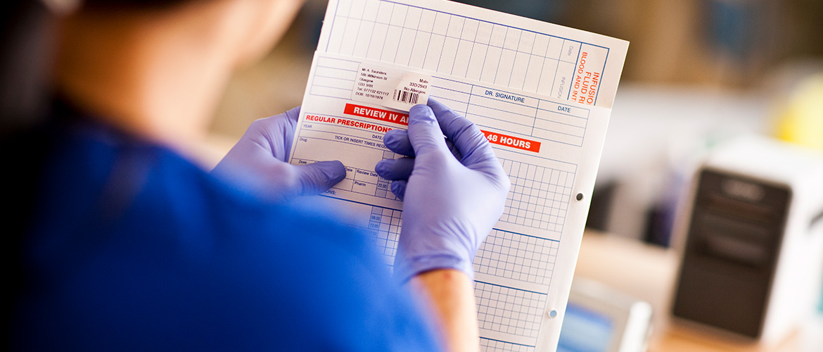 Krankenpflegerin klebt Barcode-Etikett auf Patientenakte. Etikettendrucker im Hintergrund.